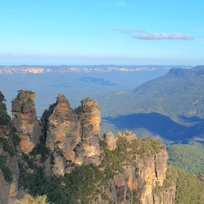 Three towering rock formations, known as the Three Sisters, surrounded by forested valleys and distant mountain ranges under a clear blue sky.