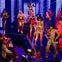 Performers in vibrant costumes dance and sing on a stage surrounded by an audience under colorful lights.