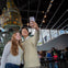 Two people taking a selfie in front of a decorative structure indoors with other visitors visible in the background.