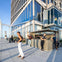 A rooftop terrace with people enjoying the space, a modern building with glass walls, and a bar counter under sunlight.