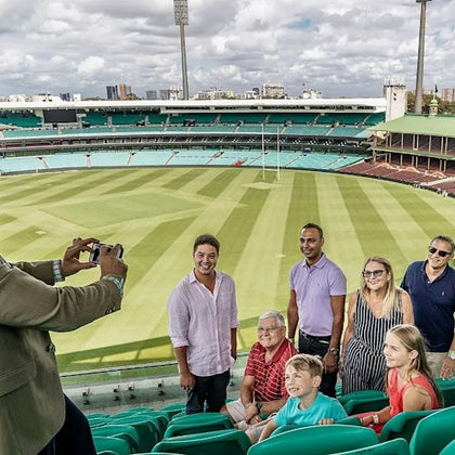 A group of people posing for a photo in a stadium with green seating and a grass field under a cloudy sky.