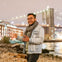 A person in a light denim jacket and scarf gestures while standing near a river with a lit bridge in the background.