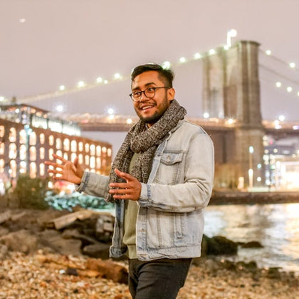 A person in a light denim jacket and scarf gestures while standing near a river with a lit bridge in the background.