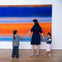 A woman holding hands with two children in front of a large abstract painting featuring vibrant blue and orange hues.