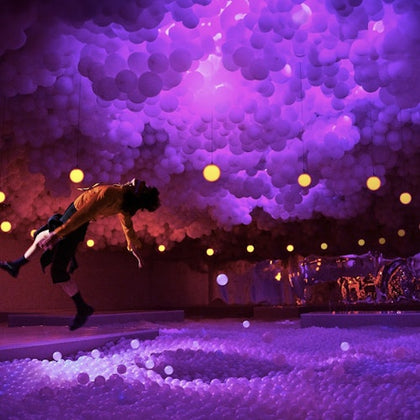A person is suspended mid-air in a room with purple lighting, surrounded by hanging balloons and illuminated spheres.