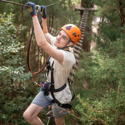 A person wearing a harness and orange helmet climbs a rope in a treetop adventure course surrounded by greenery.