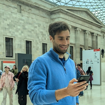 A man in a blue sweater looks at his phone in a large indoor space with architectural details and other people around.