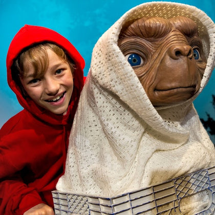 Smiling child in a red hoodie poses with a figure resembling an extraterrestrial wrapped in a white blanket against a blue background.