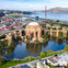 Aerial view of the Palace of Fine Arts in San Francisco, with a pond, nearby buildings, and the Golden Gate Bridge in the distance.