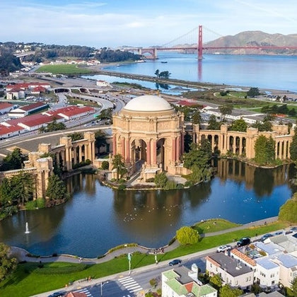 Aerial view of the Palace of Fine Arts in San Francisco, with a pond, nearby buildings, and the Golden Gate Bridge in the distance.