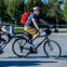 A person wearing a helmet and a red backpack is riding a bike with a child on a connected trailer bike in a park.