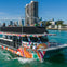 A colorful boat named "Fiesta 1" on turquoise water with people on board, city buildings and palm trees in the background.