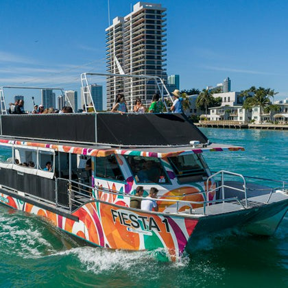 A colorful boat named "Fiesta 1" on turquoise water with people on board, city buildings and palm trees in the background.