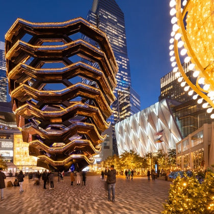 People walking near an illuminated, honeycomb-like structure and decorated tree in an urban plaza at dusk.
