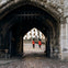 Two guards in red uniforms and black bearskin hats stand under a stone archway, with a historic building visible in the background.