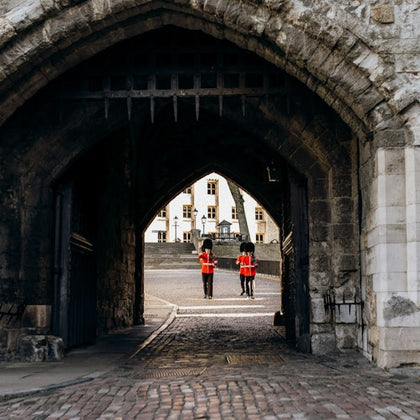 Two guards in red uniforms and black bearskin hats stand under a stone archway, with a historic building visible in the background.