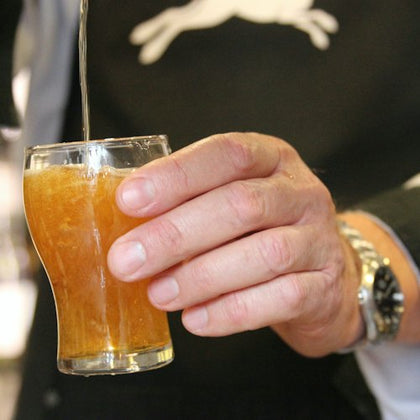 A hand holding a small glass of amber-colored liquid being poured, with a watch visible on the wrist of the hand.