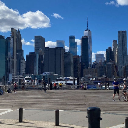 Skyline of tall buildings under a blue sky with clouds, people walking and biking on a boardwalk, and boats docked by the water.