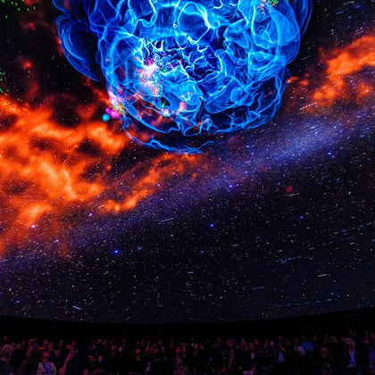 A planetarium dome showing a cosmic scene with a blue nebula, orange gas clouds, and a starry background above an audience.