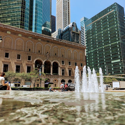 An urban plaza featuring a fountain, people sitting nearby, and tall buildings in the background under a clear sky.