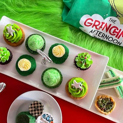A festive dessert spread featuring green cupcakes, sandwiches, and a 'Grinch Afternoon' branded cloth on a green surface.