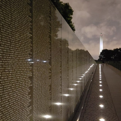A reflective wall with engraved names, illuminated by ground lights, leading to the Washington Monument at night.