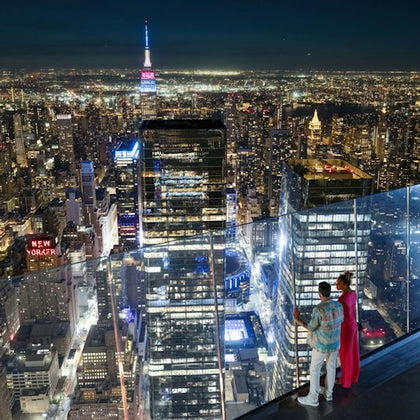 Two people stand on a glass observation deck overlooking a nighttime cityscape with skyscrapers and illuminated buildings.