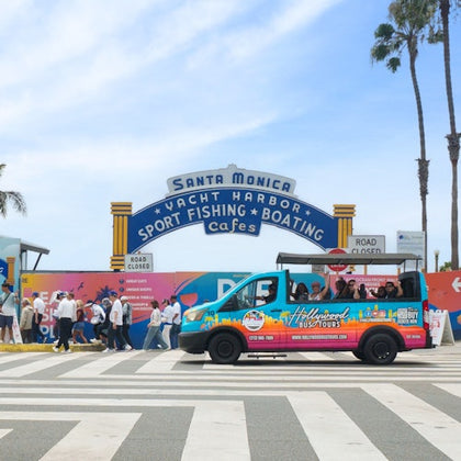 A colorful tour van parked by the Santa Monica Pier sign, with pedestrians crossing and palm trees in the background.