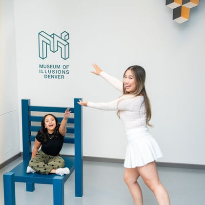 Two people pose with a large chair illusion, one sitting on it, the other pointing at the Museum of Illusions Denver sign.