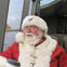 An elderly man dressed in a Santa outfit with a white beard, wearing glasses, sits by a window holding a coffee cup on a boat.
