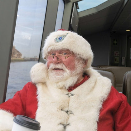 An elderly man dressed in a Santa outfit with a white beard, wearing glasses, sits by a window holding a coffee cup on a boat.