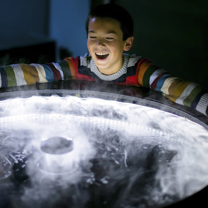 A smiling child interacts with a circular display emitting light and mist, wearing a colorful striped sweater.