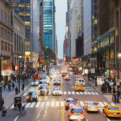 A busy city street with cars, taxis, and pedestrians. Buildings line both sides, and the scene is lively during dusk.