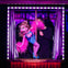 A colorful puppet in a blue outfit and pink feather boa is operated by a person dressed in black on a small stage with curtains.