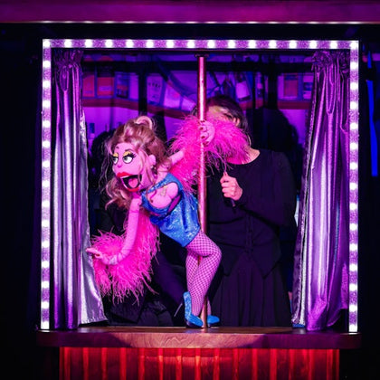 A colorful puppet in a blue outfit and pink feather boa is operated by a person dressed in black on a small stage with curtains.