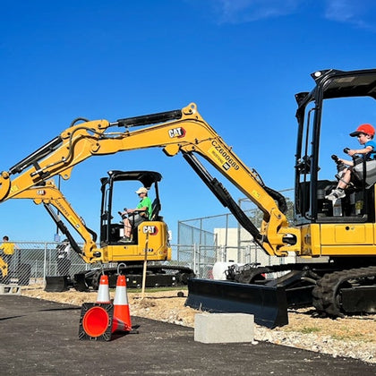 Children wearing helmets sitting on small yellow excavators in an outdoor area with traffic cones and a clear sky.