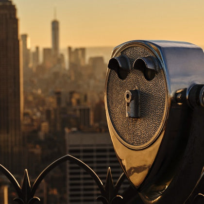 A coin-operated binocular viewer overlooking a city skyline during a golden sunset, with skyscrapers in the background.