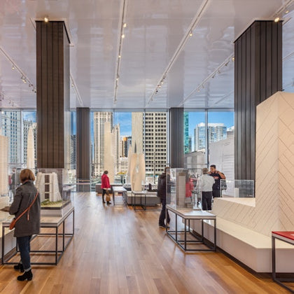 People observing architectural models in a modern, spacious room with large windows overlooking a cityscape.