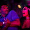A man in a blue shirt reacts with surprise as a woman in makeup and jewelry gestures animatedly under purple lighting.