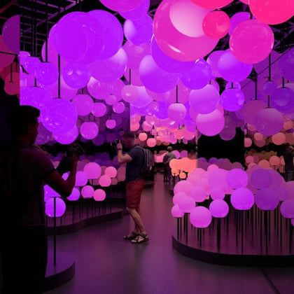 People explore a dimly lit art installation with glowing pink and purple spherical lights on thin black stands.