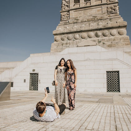 Two women pose for a photo in front of a stone monument while a person lies on the ground taking their picture with a smartphone.