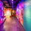 A brightly lit hallway with vibrant, psychedelic murals and text panels on the walls, leading to an orange door at the end.