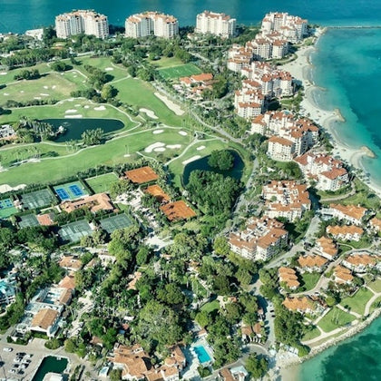 Aerial view of a coastal area with residential buildings, golf courses, tennis courts, lush greenery, and turquoise waters.