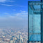 A glass observation deck juts out from a high-rise building, overlooking a cityscape with clear skies.