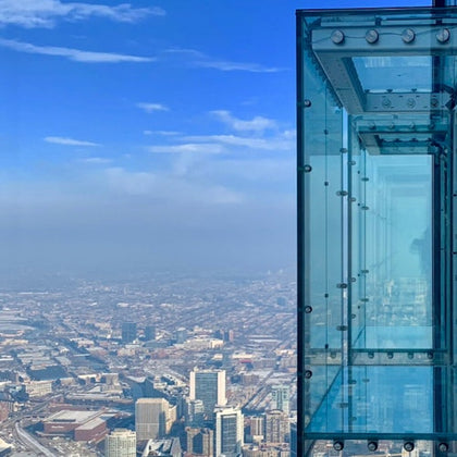 A glass observation deck juts out from a high-rise building, overlooking a cityscape with clear skies.