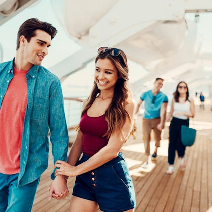 Two people smiling and holding hands while walking on a sunny deck, with another couple in the background.