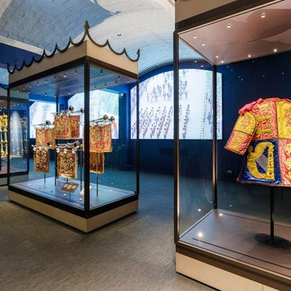 Exhibit with ornate, historical garments in glass cases; screen displaying a parade in the background.