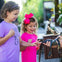 Two young girls interact with a small alligator held by a person, outdoors in a sunny setting.