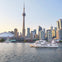 A city skyline with a prominent tower and several buildings by the waterfront, with boats on the water in the foreground.