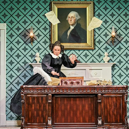 A woman in period costume stands behind a desk with papers flying, in a room with a portrait of George Washington on the wall.